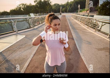 Beautiful confident sportswoman, female athlete, jogger, runner standing on a treadmill and putting on earphones, getting ready to run over the city b Stock Photo