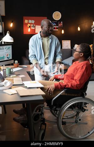 African woman sitting on wheelchair and discussing the work with her colleague at office Stock Photo