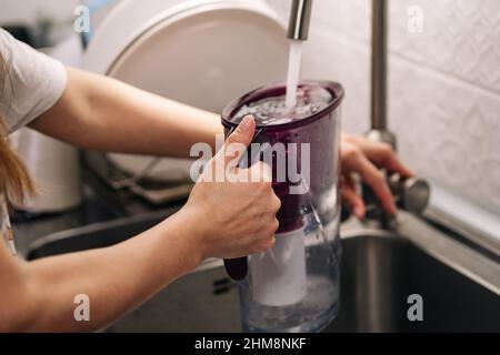 Woman pouring water from faucet into water filter jug at the kitchen. Healthy lifestyle. Woman filling water. Stock Photo