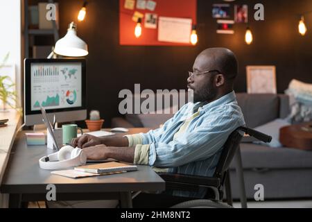 African man sitting at the table on wheelchair andworking online on laptop in the room at home Stock Photo