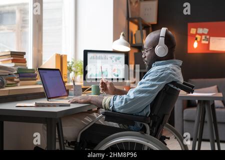 African man with disability sitting on wheelchair in headphones and using laptop during study at the table at home Stock Photo