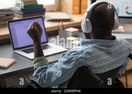 Rear view of African man in wireless headphones sitting on wheelchair and studying online on laptop at home Stock Photo