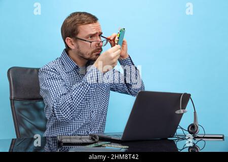 Portrait of man in glasses and checkered shirt attentively looking at motherboard isolated over blue background Stock Photo