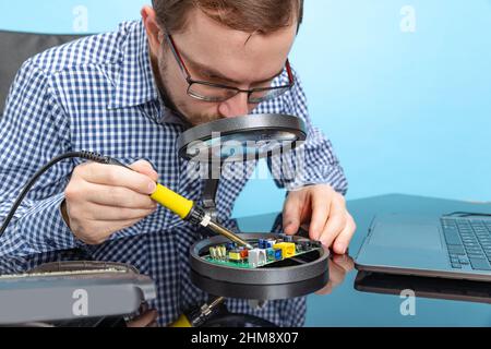 Portrait of concentrated man, computer geek looking in magnifying glass on motherboard isolated over blue background Stock Photo