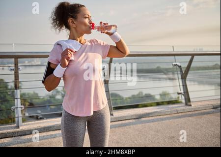 Happy sportswoman with aesthetic healthy body in sportswear drinks water after outdoor workout, stands on a bridge treadmill and enjoys beautiful sunn Stock Photo