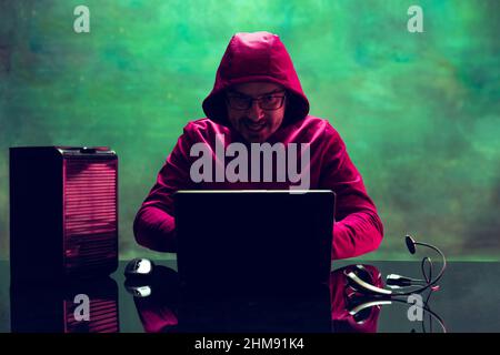 Portrait of man, computer geek, hacker working with laptop, breaking security system isolated over dark green background in neon Stock Photo