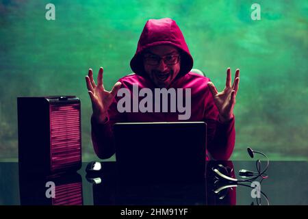 Portrait of man, computer geek, hacker working with laptop, breaking security system isolated over dark green background in neon Stock Photo