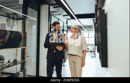 Diverse businesspeople sharing a laugh during a discussion in an office. Two happy business colleagues using a digital tablet while walking together i Stock Photo
