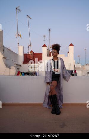 African American woman in stylish clothes with sunglasses and leaning on barrier against houses on terrace in evening, Cadiz,Spain Stock Photo