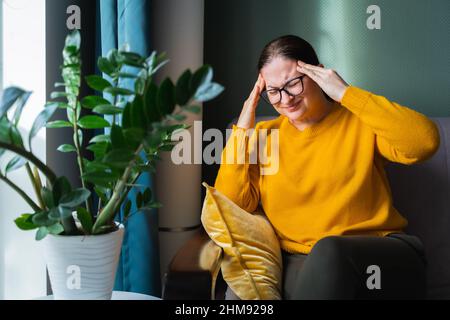 Portrait of tired woman glasses, yellow sweater, sitting on sofa near plant. Holding forehead, having headache. Migraine Stock Photo