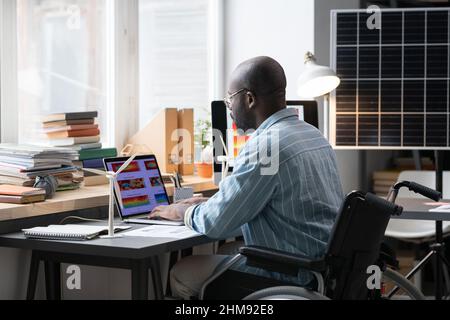 Rear view of African architect sitting in wheelchair at his workplace and typing on laptop, he developing new project online Stock Photo