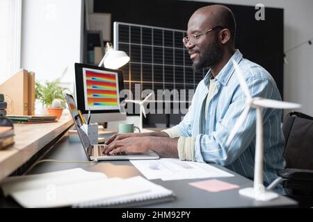African man in eyeglasses sitting at his workplace with documents and typing on laptop during his work at office Stock Photo