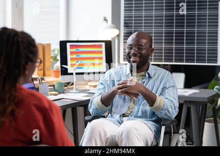 African man sitting on wheelchair and talking to his colleague during break at office Stock Photo