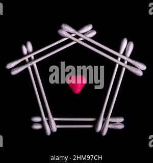 Some cotton buds are arranged on a shiny black surface with the shape of a house and a red heart in the center Stock Photo