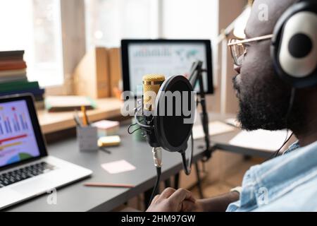 Close-up of African man in headphones speaking to the microphone while sitting at the table and using laptop during his podcast Stock Photo