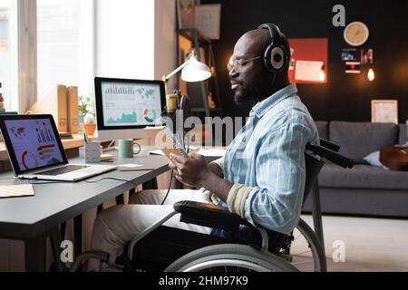 African young man in headphones sitting on wheelchair and reading something in his notebook during his online study at the table Stock Photo