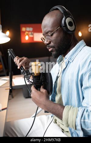 African young man in headphones sitting in front of the microphone and preparing for online broadcasting Stock Photo
