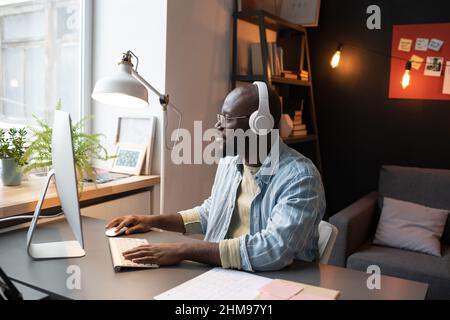 African man in wireless headphones sitting at his workplace in front of the computer monitor and working Stock Photo