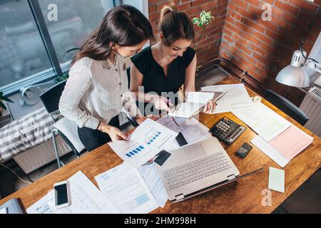 Top view of two female colleagues and desk covered with papers and documents. Businesswomen studying the data working on new sales strategy. Stock Photo