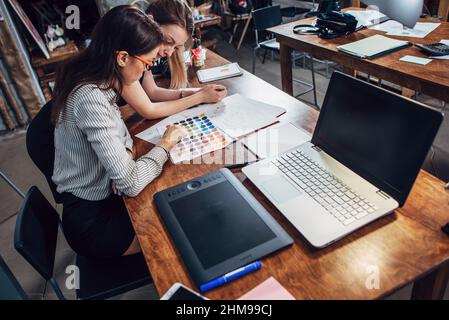 Two female architects working together using color swatches sitting at desk with laptop, graphic tablet in design studio. Stock Photo