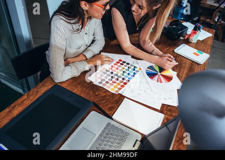 Two female architects working together using color swatches sitting at desk with laptop, graphic tablet in design studio. Stock Photo