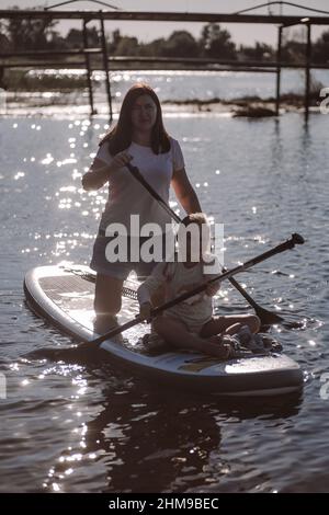 Female child with mother sup boarding with oars in hands smiling and looking at camera on lake shining from sun. Active lifestyle. Teaching children Stock Photo