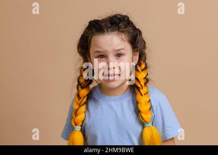 Sad Caucasian young girl crying with open mouth and wet eyes with kanekalon braids of yellow color on head wearing light blue t-shirt on beige Stock Photo