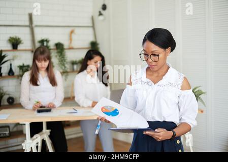 Smiling young dark-skinned woman stands in a modern office with her colleagues in the background. Female designer with coworkers in conference room. Stock Photo