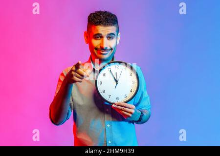 Portrait of smiling man in shirt pointing finger at camera holding big wall clock in hand, time to action, motivation. Indoor studio shot isolated on colorful neon light background. Stock Photo