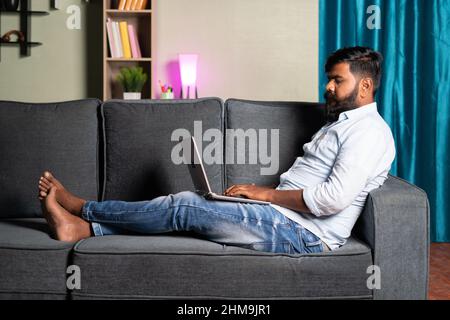 Wide angle shot of relaxed young man busy using laptop while sitting on safa at home - concept of work from home, new normal employee or job lifestyle Stock Photo
