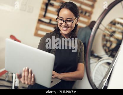Emails are how I stay in touch with clients. Shot of an attractive young woman standing alone in her bicycle shop and using her laptop. Stock Photo