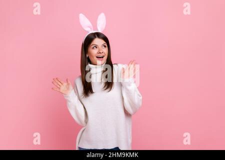 Portrait of excited young adult brunette woman with bunny ears, looking away, childish behavior, wearing white casual style sweater. Indoor studio shot isolated on pink background. Stock Photo