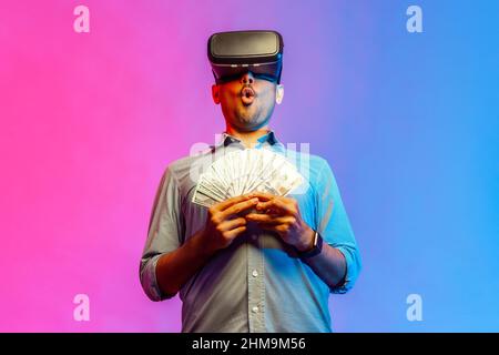 Portrait of amzed man in shirt to get money, holding dollars, illusion of rich millionaire, playing virtual reality game. Indoor studio shot isolated on colorful neon light background. Stock Photo