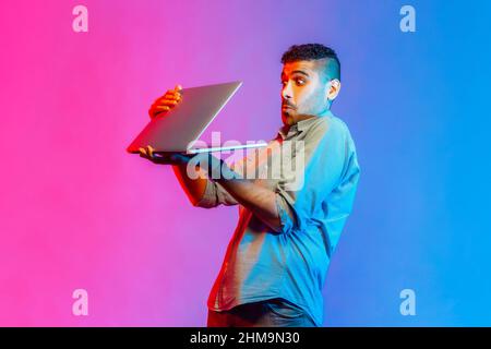 Portrait of young adult man in shirt holding laptop in hands, sees shocking scared content, looking at display with big eyes. Indoor studio shot isolated on colorful neon light background. Stock Photo