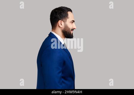 Side view portrait of cheerful bearded man with smile, standing and looking at camera, expressing positive emotions, wearing official style suit. Indoor studio shot isolated on gray background. Stock Photo