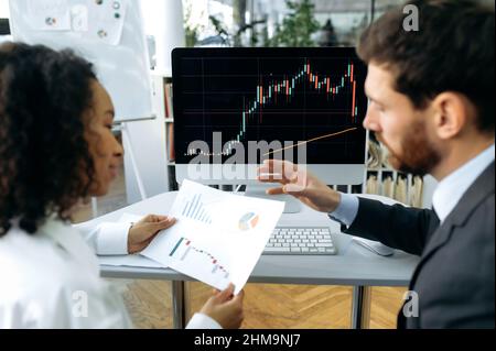 Focused multiracial trading colleagues, Caucasian man and African American woman, crypto traders, using a computer, looking at charts, analyzing the crypto market, planning a strategy for good profit Stock Photo