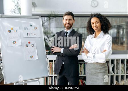 Successful intelligent multiracial colleagues, caucasian man and african american woman, in formal clothes, are standing with arms crossed in modern office near whiteboard, looking at camera and smile Stock Photo