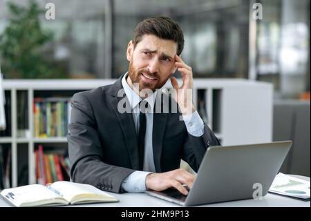 Sad overworked caucasian male business person, manager or company owner, in modern office, suffering from headache, migraine, rubs temple, feels stressed, tired from overtime work, stressful situation Stock Photo