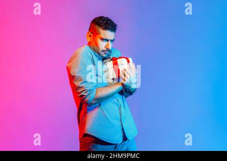 Portrait of man in shirt holding present box in hands, being greedy to share gifts, looking at camera with frowning face. Indoor studio shot isolated on colorful neon light background. Stock Photo