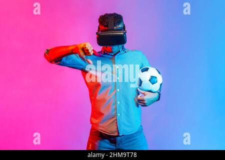 Portrait of man in shirt wearing virtual reality glasses, standing with soccer ball, showing thumb down, dislike football match. Indoor studio shot isolated on colorful neon light background. Stock Photo