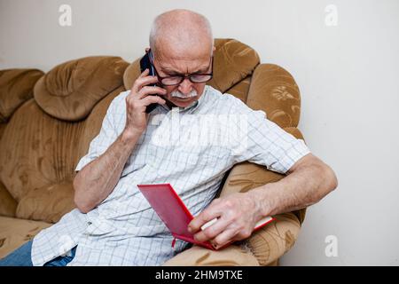 elderly man speaks on his cell phone checking data in a paper notebook. old businessman plans purchases and calls store. Senior citizen plans spending Stock Photo