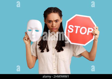 Serious woman with black dreadlocks holding white mask and red stop sign, looking at camera, do not change personality, wearing white shirt. Indoor studio shot isolated on blue background.