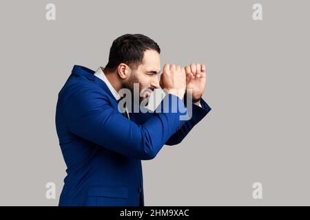 Side view of concentrated bearded businessman looking through monocular, having serious facial expression, wearing official style suit. Indoor studio shot isolated on gray background. Stock Photo
