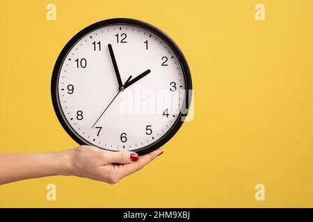 Profile side view closeup of woman hand holding big wall clock, deadline, time to go. Indoor studio shot isolated on yellow background. Stock Photo