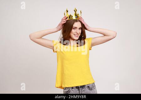 I am queen. Portrait of happy curly-haired teenager girl putting crown on head and smiling, self confidence, self-motivation and dreams to be best. Indoor studio shot isolated on gray background. Stock Photo