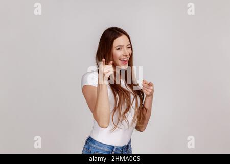 Portrait of beautiful happy woman pointing finger and winking to camera, choosing winner, gesturing we need you, wearing white T-shirt. Indoor studio shot isolated on gray background. Stock Photo