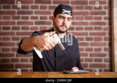 Focused male cook burning slice of tuna on rice with gas burner while preparing traditional sushi during work in restaurant Stock Photo
