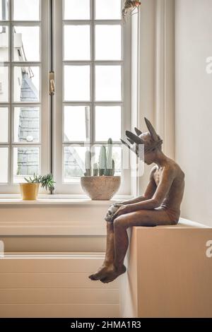 Statue of man in crown placed on white cabinet near potted green plants at window in light room at home Stock Photo