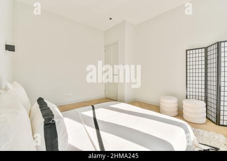 Comfortable bed with white cushions and bedclothes placed on rug in light spacious bedroom with doors and poufs at home Stock Photo