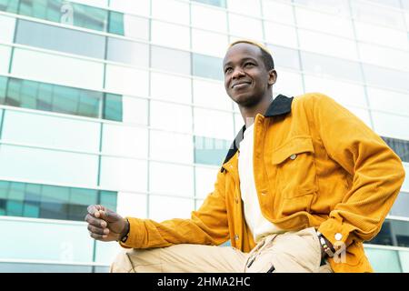 Peaceful smiling African American male hipster with backpack chilling sitting in park and drinking refreshing beverage to go Stock Photo
